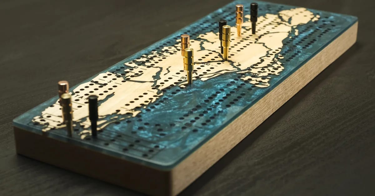 Planche de cribbage dont le dessus reprend la carte de la Gaspésie découpée dans l'érable, avec une résine turquoise pour la mer et des fiches de métal