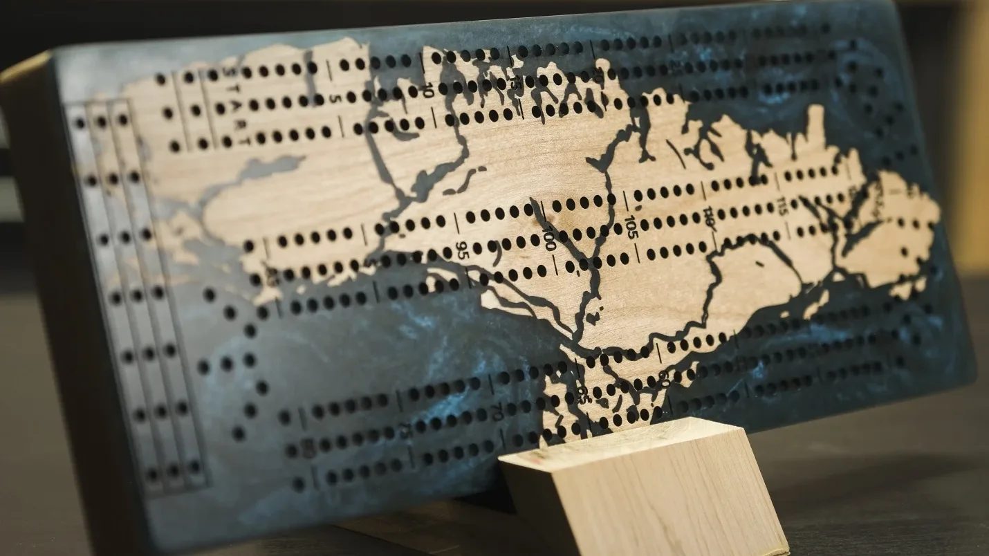 Planche de cribbage en érable et résine bleu ardoise, posée sur un support en bois, avec les rangées de trous et les points gravés