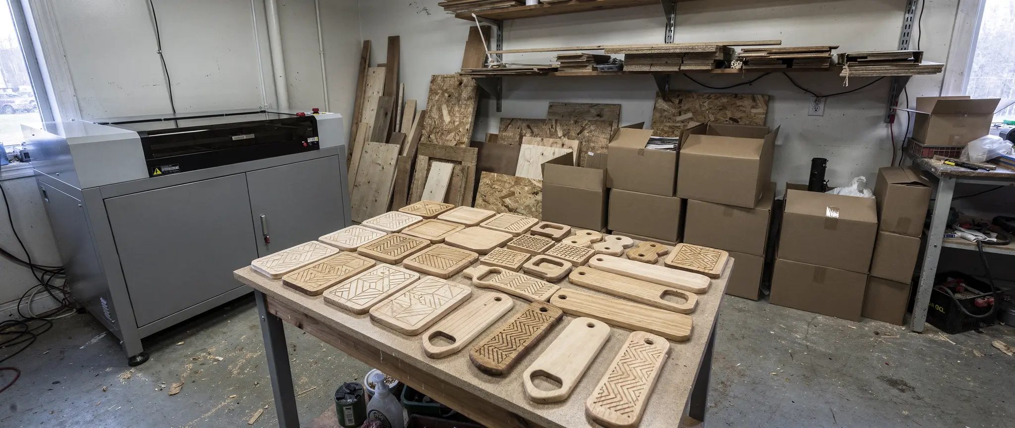 Illustration de référence : objets promotionnels personnalisés en bois gravé, série de sous-verres, étiquettes et décapsuleurs sur une table près d'une machine laser, avec des boîtes de carton prêtes pour l'expédition