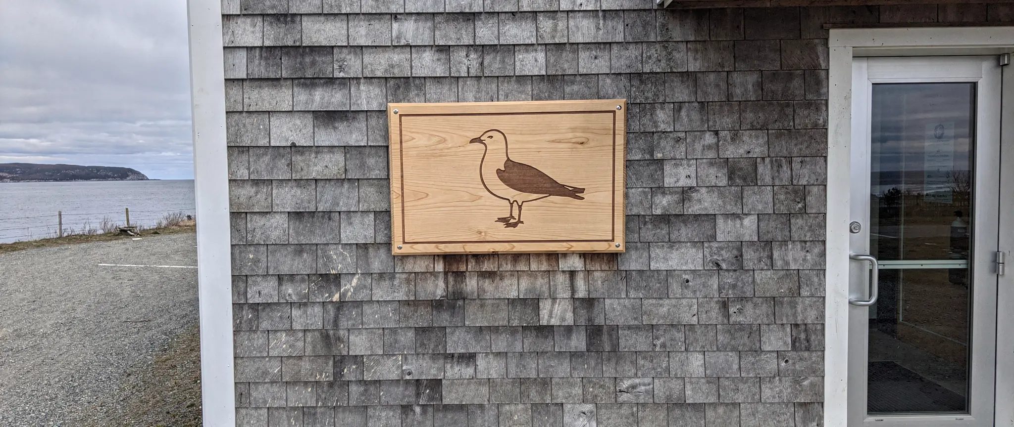 Reference illustration: custom engraved commemorative plaque in birch with a gull and a thin border, mounted on the cedar-shingle wall of a small community building facing the Baie des Chaleurs
