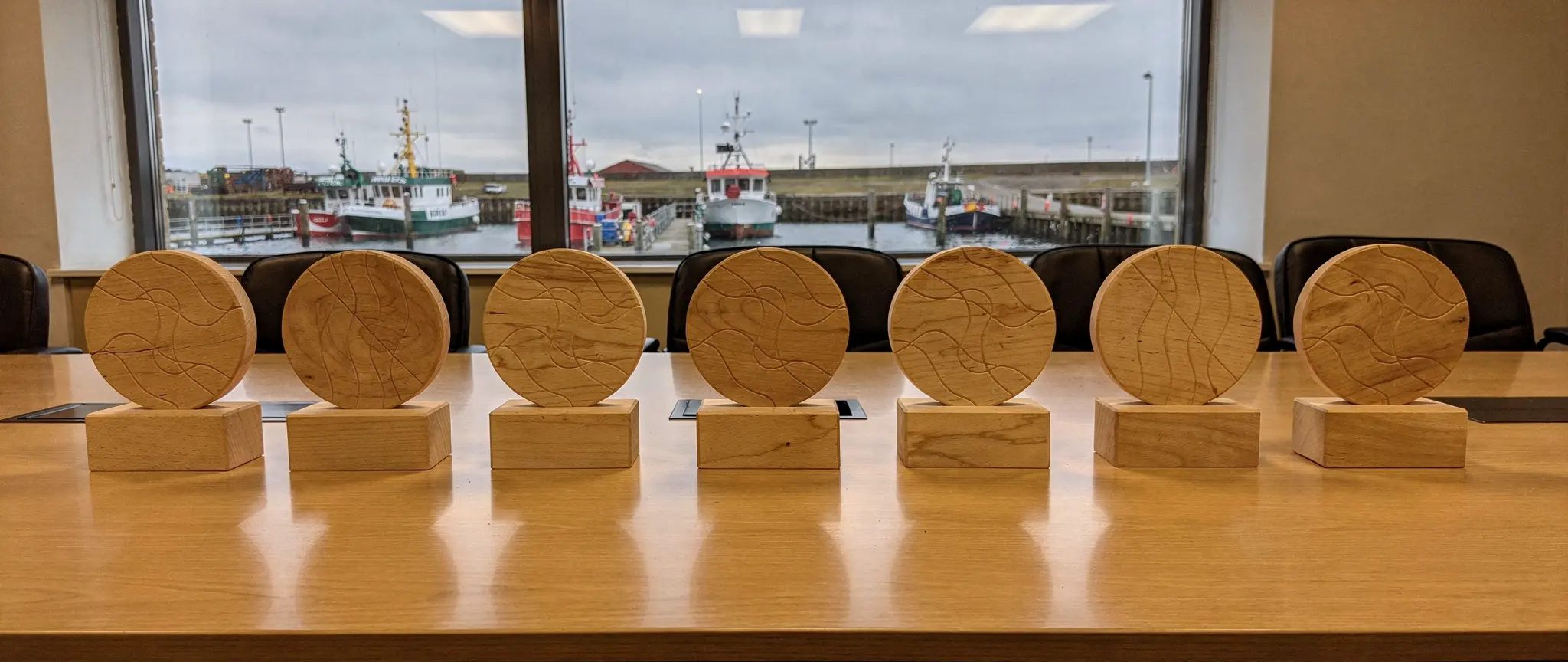 Illustration de référence : trophées personnalisés en bois pour une entreprise, sept pièces identiques (disque gravé de vagues sur un socle) alignées sur une table de salle de réunion devant une fenêtre sur le port, avant une remise annuelle